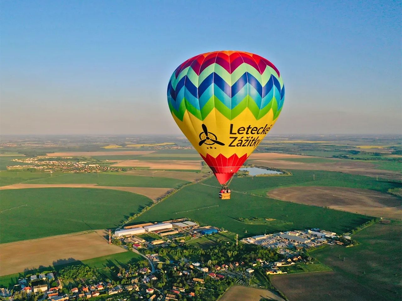 Let balónom Letecké Zážitky. Farebný balón letí nad zelenými poliami a dedinkami. Pokojný let balónom s výhľadom, ktorý berie dych.