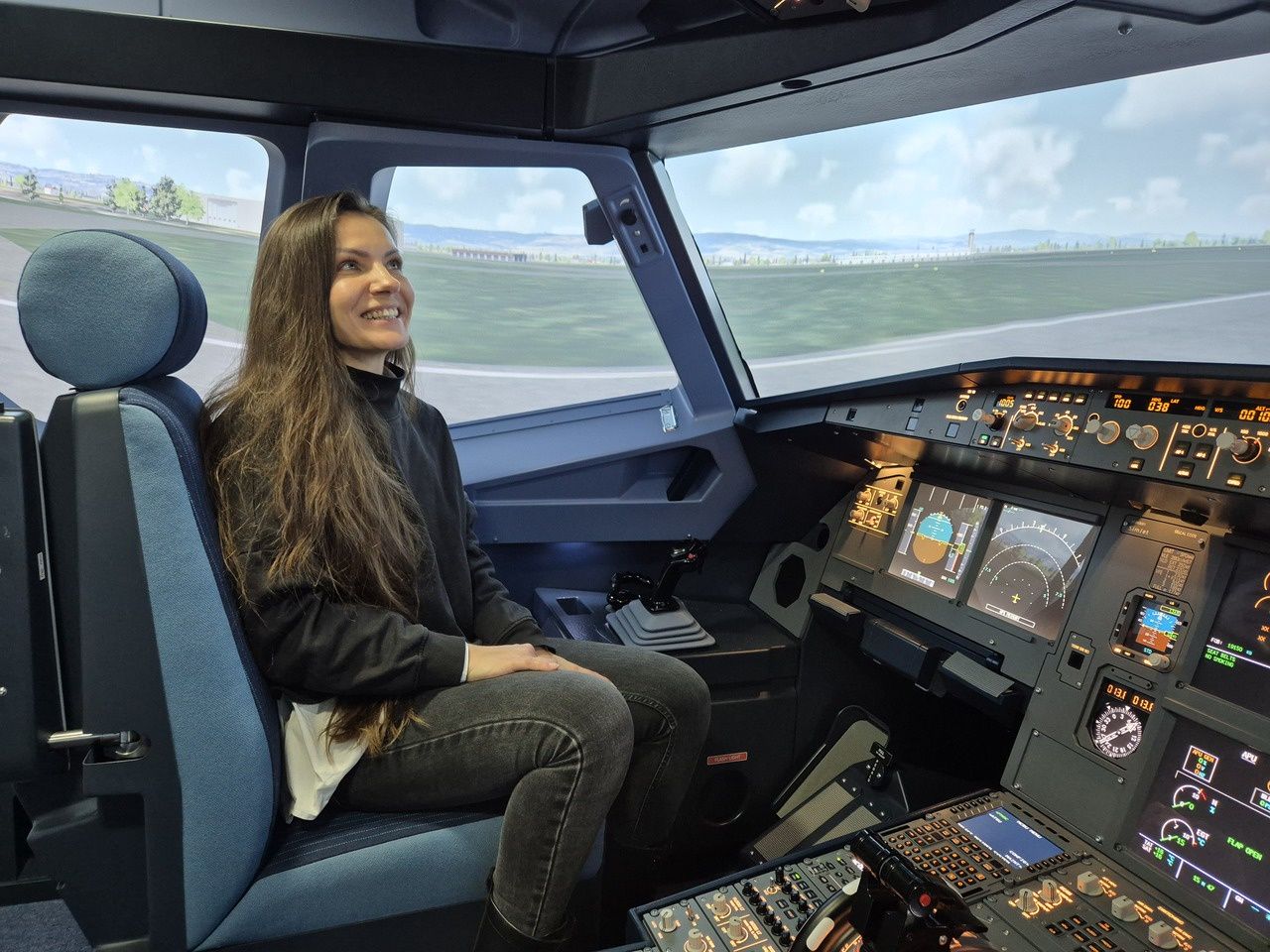Žena sedí v kokpite leteckého simulátora Airbus a s úsmevom sa pozerá cez okno na virtuálnu pristávaciu dráhu.